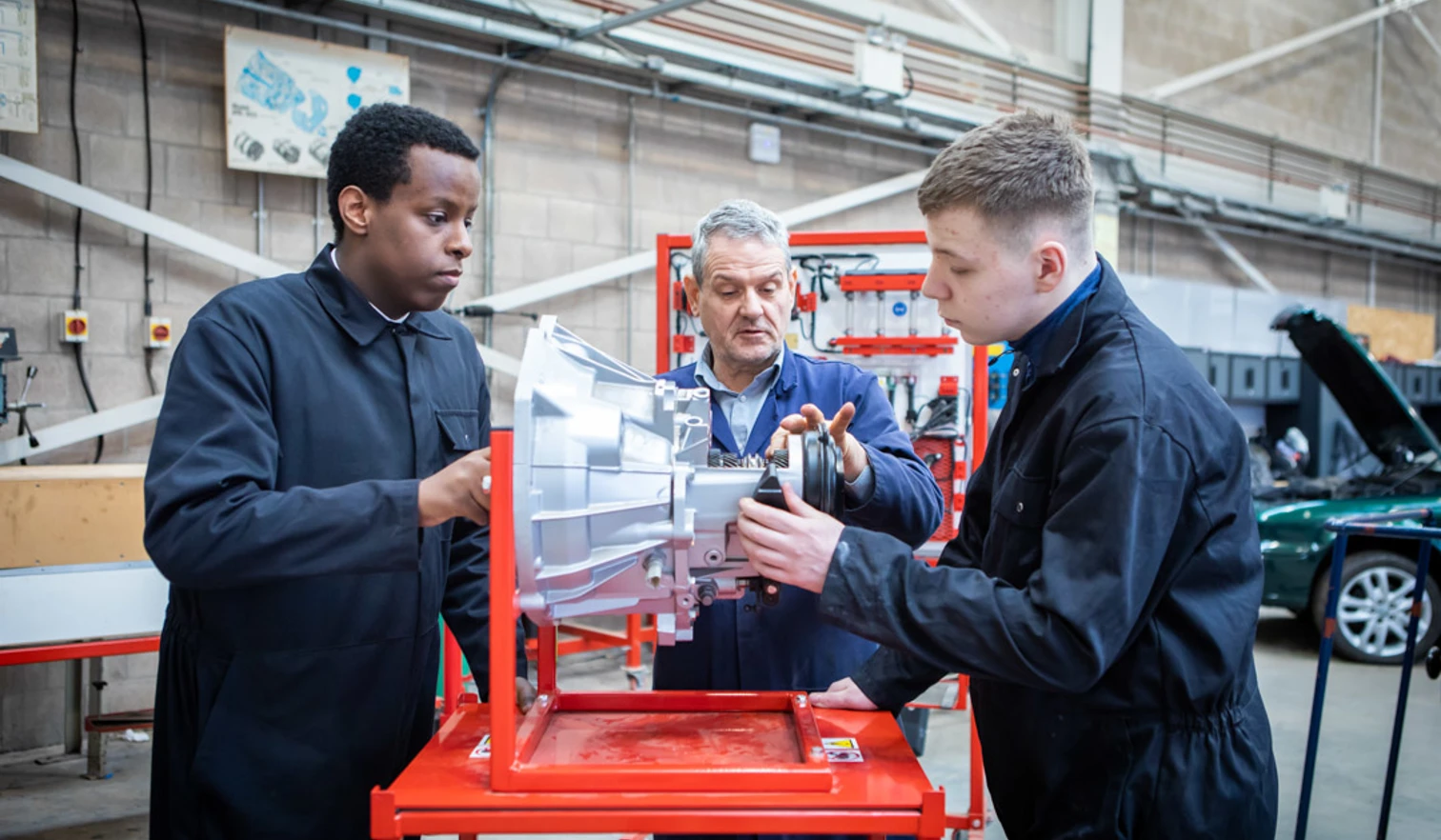 Automotive instructor guiding two students in overalls as they work on a mechanical component in a workshop. Automotive instructor guiding two students in overalls as they work on a mechanical component in a workshop.